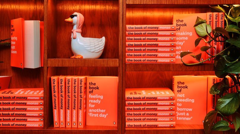 Orange books on a bookshelf. The book's title is "The book of Money" the author is Monzo. There is also a duck statue. The duck has sunglasses and a scarf over its head.